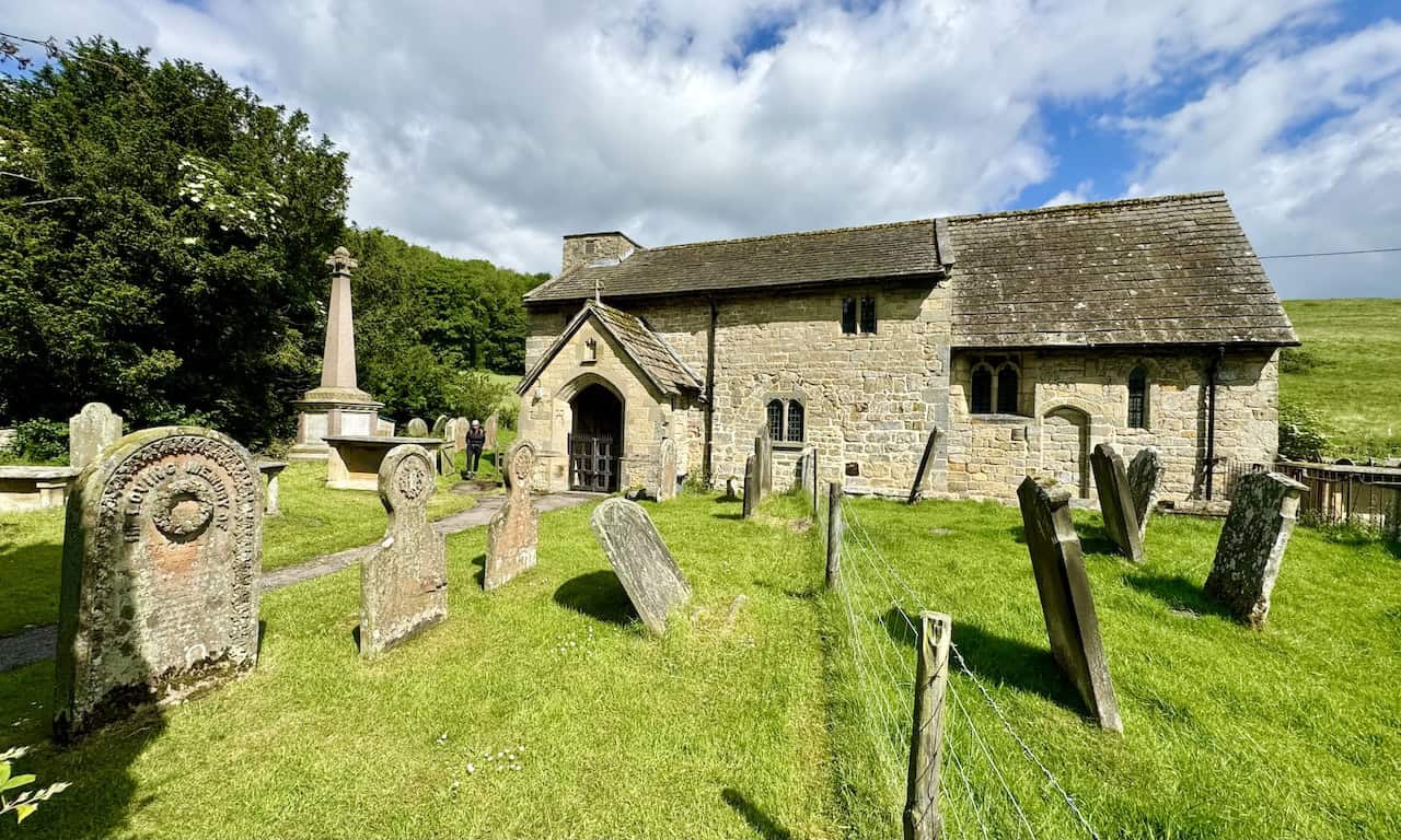 The churchyard of St Hilda’s hosts a collection of gravestones and memorials, including a prominent tall obelisk war memorial to the left of the porch entrance, creating a picturesque setting.