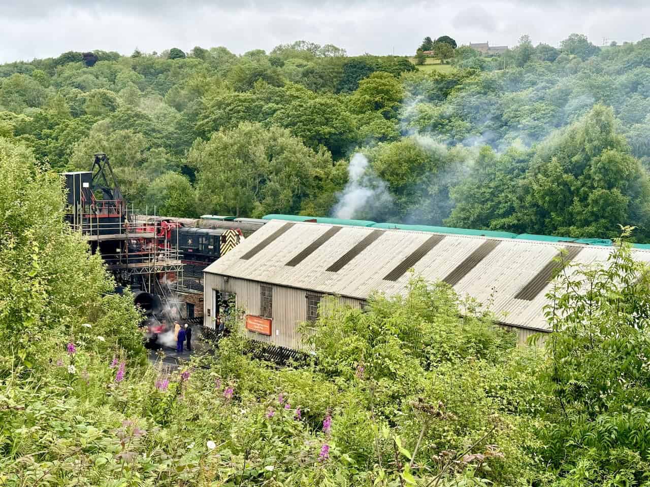 Viewing area with a bench at the top of some steps. Below are the engine sheds and works of the North Yorkshire Moors Railway, set within the lush green landscape.