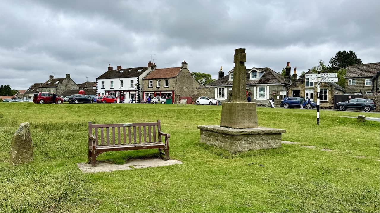 Goathland village green with a wooden bench and memorial cross, surrounded by shops and cafes on the Grosmont to Goathland walk.