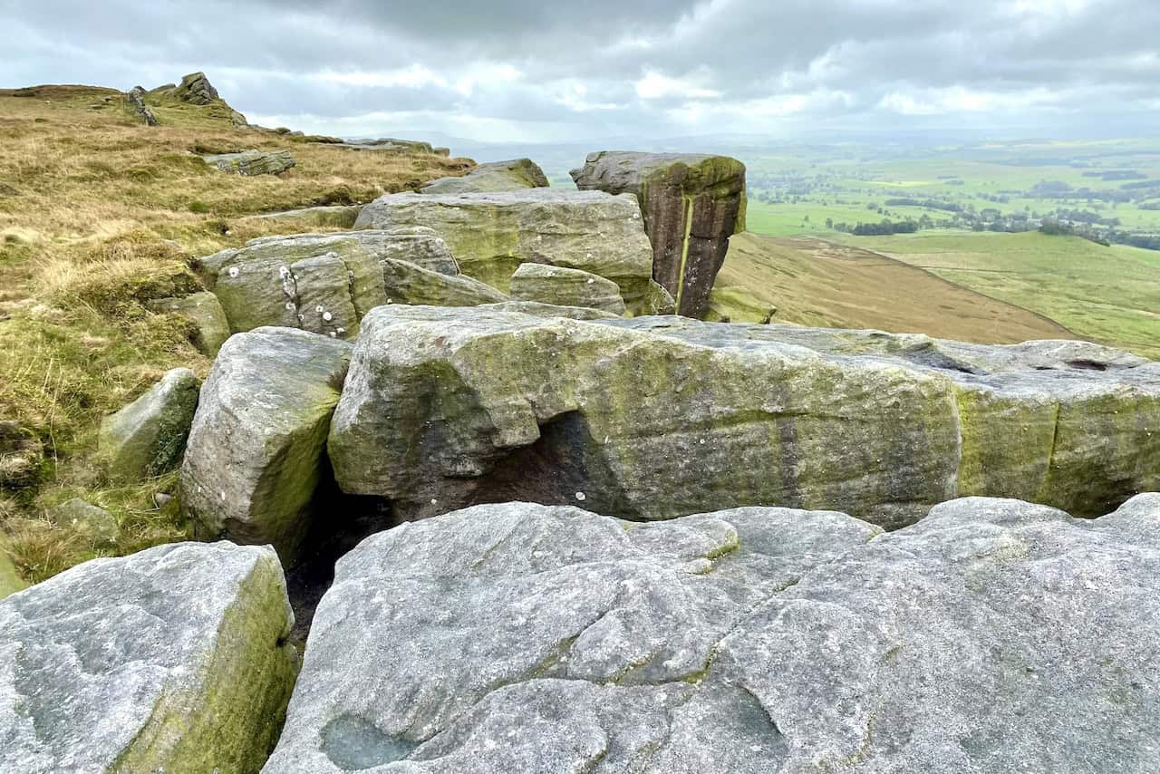 Rolling Gate Crags, a rugged outcrop of large, weathered rocks on the hillside, with a wide green valley, Cracoe village, and Swinden Quarry visible in the background.