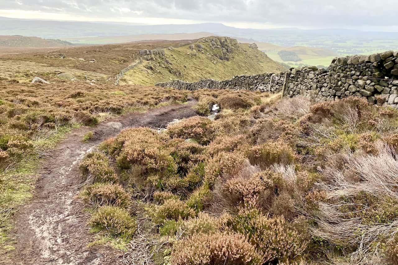 Following the dry stone wall towards the cross on Rylstone Fell, with heather and rough moorland terrain dotted with boulders.