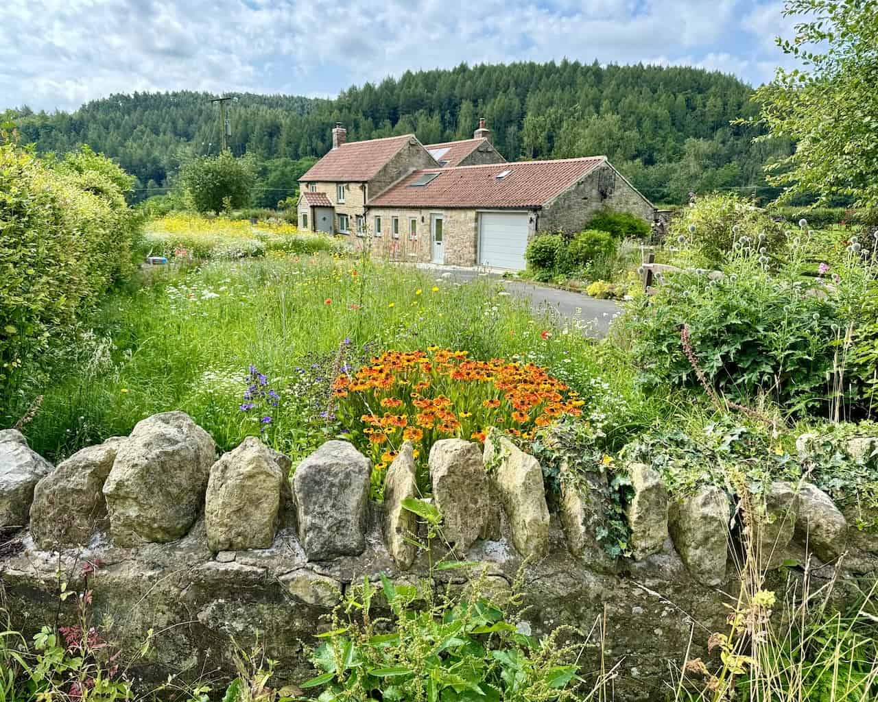 A picturesque scene of a stone cottage with a red-tiled roof, surrounded by flowers and trees.
