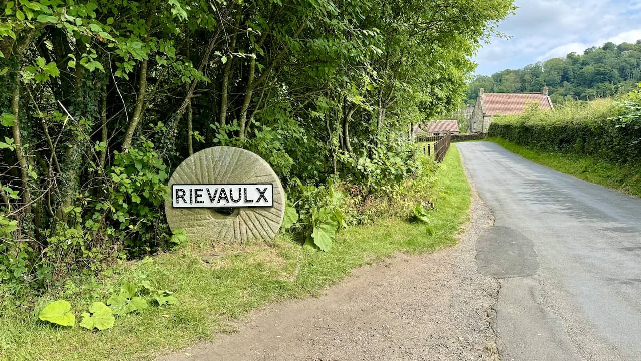 A narrow road leading into the village of Rievaulx, running parallel to the River Rye.
