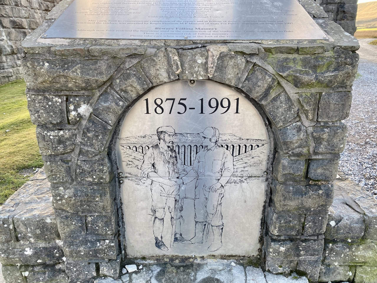 Silver plaque on a stone statue near Ribblehead Viaduct, depicting a 19th-century navvy shaking hands with a 20th-century engineer.